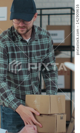 Vertical video. The courier comes to pick up the package for the customer. Female manager using laptop computer preparing package for postal delivery. Vertical video. The courier comes to pick up the package for the customer. Female manager using laptop computer preparing package for postal delivery. 142909616