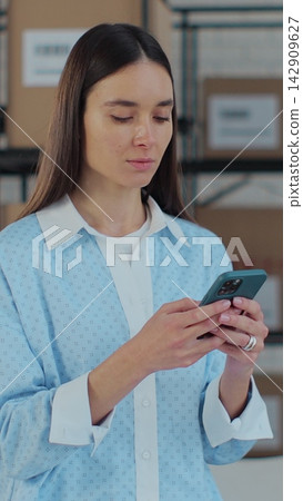 Vertical video. Portrait of a woman using a smartphone at work in a warehouse. A woman connects to customer support via the Internet. Work manager in an online store. Vertical video. Portrait of a woman using a smartphone at work in a warehouse. A woman connects to customer support via the Internet. Work manager in an online store. 142909627