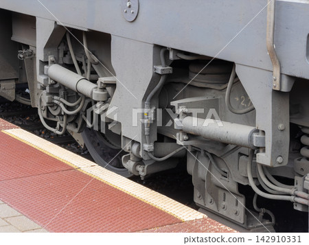 A close view of the heavy steel bogie under a passenger train parked at a station platform, highlighting mechanical engineering components of modern public transport infrastructure A close view of the heavy steel bogie under a passenger train parked at a station platform, highlighting mechanical engineering components of modern public transport infrastructure 142910331