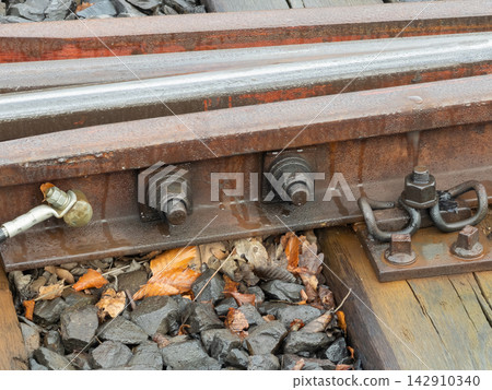 A close up view of heavy metal train tracks showing rusty bolts and steel plates on wooden sleepers with scattered autumn leaves which highlights railway infrastructure maintenance work A close up view of heavy metal train tracks showing rusty bolts and steel plates on wooden sleepers with scattered autumn leaves which highlights railway infrastructure maintenance work 142910340