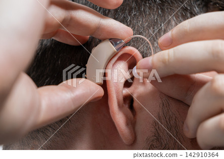 A man carefully inserts a discreet beige behind the ear hearing aid into his ear canal to improve auditory function for daily communication in a healthcare and personal wellness context A man carefully inserts a discreet beige behind the ear hearing aid into his ear canal to improve auditory function for daily communication in a healthcare and personal wellness context 142910364