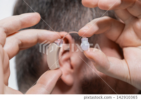 A man places a modern beige hearing aid behind his ear, demonstrating medical care, accessibility, and improved auditory health for individuals dealing with everyday communication loss A man places a modern beige hearing aid behind his ear, demonstrating medical care, accessibility, and improved auditory health for individuals dealing with everyday communication loss 142910365