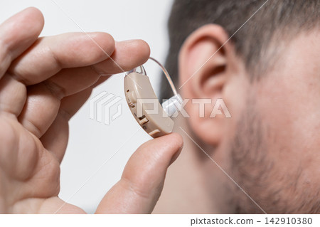 A person holds a modern beige hearing aid near their ear to illustrate medical sound amplification technology for improving daily communication and restoring auditory health for seniors A person holds a modern beige hearing aid near their ear to illustrate medical sound amplification technology for improving daily communication and restoring auditory health for seniors 142910380