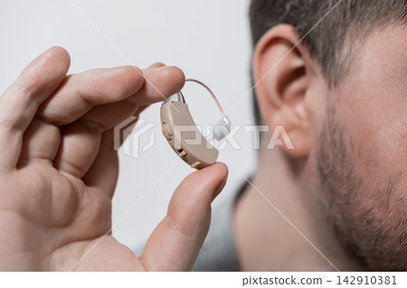 A man holds a modern behind-the-ear hearing aid between his fingers to highlight medical technology and assistive equipment for individuals with hearing loss in daily life A man holds a modern behind-the-ear hearing aid between his fingers to highlight medical technology and assistive equipment for individuals with hearing loss in daily life 142910381