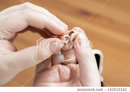 Hands holding a beige hearing aid device for maintenance, highlighting the delicate process of adjusting medical equipment to improve auditory accessibility and daily personal care Hands holding a beige hearing aid device for maintenance, highlighting the delicate process of adjusting medical equipment to improve auditory accessibility and daily personal care 142910382