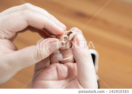 A person holds a small hearing aid device, carefully inspecting or adjusting the plastic casing component to ensure proper function and maintenance for improved daily sound reception A person holds a small hearing aid device, carefully inspecting or adjusting the plastic casing component to ensure proper function and maintenance for improved daily sound reception 142910383