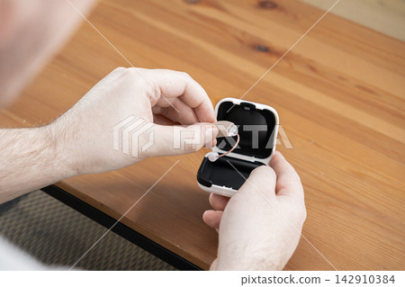 A man handles a modern hearing aid device near an open charging case on a wooden table, representing accessible medical technology for better communication and daily audiology wellness A man handles a modern hearing aid device near an open charging case on a wooden table, representing accessible medical technology for better communication and daily audiology wellness 142910384