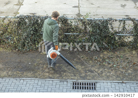 A worker uses a leaf blower to clear debris near a fence, showcasing professional groundskeeping maintenance tasks for maintaining clean outdoor environments in urban parkland areas A worker uses a leaf blower to clear debris near a fence, showcasing professional groundskeeping maintenance tasks for maintaining clean outdoor environments in urban parkland areas 142910417