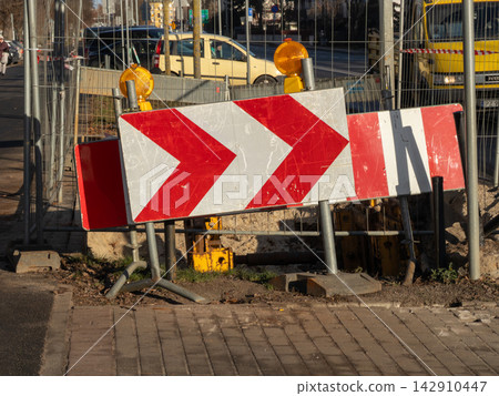 A red and white chevron road sign stands before a deep street construction trench near a metal fence, signaling a traffic diversion zone during urban infrastructure repair projects A red and white chevron road sign stands before a deep street construction trench near a metal fence, signaling a traffic diversion zone during urban infrastructure repair projects 142910447