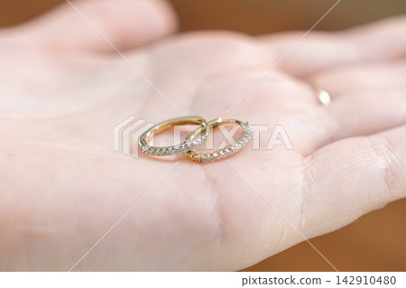 Two delicate gold hoop earrings with small sparkling diamond accents resting on a person palm, conveying luxury jewelry and high quality accessory fashion for formal elegant occasions Two delicate gold hoop earrings with small sparkling diamond accents resting on a person palm, conveying luxury jewelry and high quality accessory fashion for formal elegant occasions 142910480