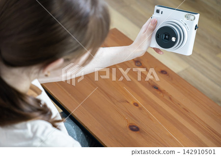 A young person holds a white instant camera to take a selfie over a wooden desk, capturing a creative moment of self expression with modern analog technology in a bright home A young person holds a white instant camera to take a selfie over a wooden desk, capturing a creative moment of self expression with modern analog technology in a bright home 142910501