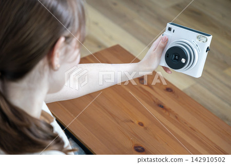 Person holding a white instant camera to take a self portrait above a wooden table, capturing a lifestyle moment with creative photography gear in a bright cozy home indoor setting Person holding a white instant camera to take a self portrait above a wooden table, capturing a lifestyle moment with creative photography gear in a bright cozy home indoor setting 142910502