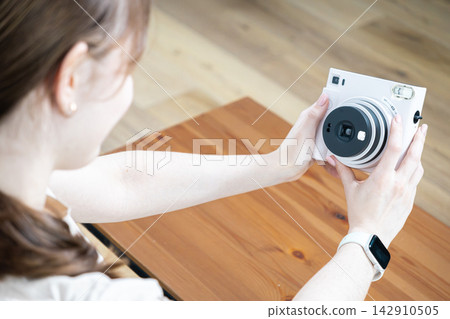 A young woman holds a white instant camera to capture a photograph at home, evoking nostalgic hobbies, creative lifestyle, and the joy of preserving memories through analogue tech A young woman holds a white instant camera to capture a photograph at home, evoking nostalgic hobbies, creative lifestyle, and the joy of preserving memories through analogue tech 142910505