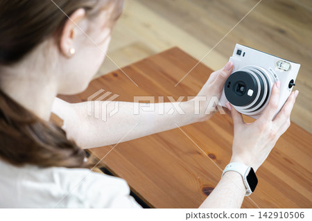 A young person captures memories using a white instant camera while sitting at a rustic wooden table, embodying a creative lifestyle and the joy of preserving precious life moments A young person captures memories using a white instant camera while sitting at a rustic wooden table, embodying a creative lifestyle and the joy of preserving precious life moments 142910506