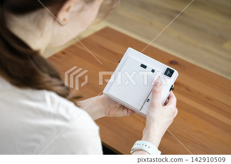 A young person holds a white instant camera over a rustic wooden table, creating a moment of creative inspiration, hobby photography, and personal leisure in a bright living space A young person holds a white instant camera over a rustic wooden table, creating a moment of creative inspiration, hobby photography, and personal leisure in a bright living space 142910509