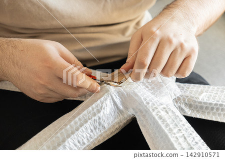 Hands using a small utility knife to cut cardboard padding on a wrapped furniture frame for delivery, demonstrating careful protective packaging in a professional moving service context Hands using a small utility knife to cut cardboard padding on a wrapped furniture frame for delivery, demonstrating careful protective packaging in a professional moving service context 142910571