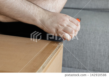A hand holding a utility knife cuts adhesive tape on a cardboard delivery box, representing the process of unboxing a package after arrival at home in a casual daily life setting A hand holding a utility knife cuts adhesive tape on a cardboard delivery box, representing the process of unboxing a package after arrival at home in a casual daily life setting 142910586