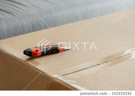 A sharp utility knife rests on a large sealed cardboard shipping box, representing the process of opening parcel deliveries or preparing for an upcoming residential moving project day A sharp utility knife rests on a large sealed cardboard shipping box, representing the process of opening parcel deliveries or preparing for an upcoming residential moving project day 142910592