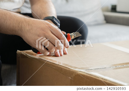 A man uses a sharp utility knife to cut adhesive tape on a brown cardboard box, reflecting the act of unpacking new deliveries inside a modern home setting during moving or arrival A man uses a sharp utility knife to cut adhesive tape on a brown cardboard box, reflecting the act of unpacking new deliveries inside a modern home setting during moving or arrival 142910593