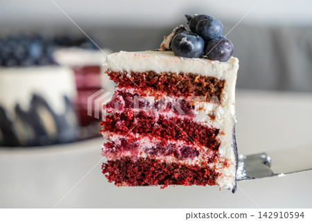 A slice of decadent red velvet cherry layer cake on a metal spatula, with fresh blueberries on top, captured in a bright kitchen for a sophisticated dessert or celebratory occasion A slice of decadent red velvet cherry layer cake on a metal spatula, with fresh blueberries on top, captured in a bright kitchen for a sophisticated dessert or celebratory occasion 142910594