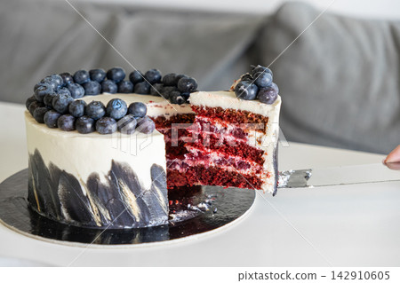 A slice of decadent red velvet cake with cream cheese frosting and fresh blueberries being lifted by a metal server on a white surface, creating an elegant mood for gourmet desserts A slice of decadent red velvet cake with cream cheese frosting and fresh blueberries being lifted by a metal server on a white surface, creating an elegant mood for gourmet desserts 142910605