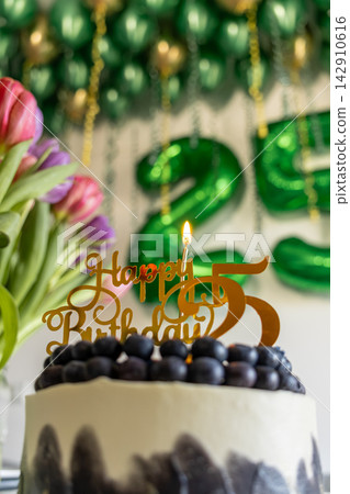 A gold cake topper with a lit candle stands on a blueberry dessert before green balloons, evoking a joyful twenty-fifth birthday celebration with vibrant flowers in a home setting A gold cake topper with a lit candle stands on a blueberry dessert before green balloons, evoking a joyful twenty-fifth birthday celebration with vibrant flowers in a home setting 142910616