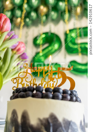 A birthday cake topped with a golden topper and a single burning candle, celebrating a milestone anniversary amidst colorful tulip flowers and green party balloons in the background A birthday cake topped with a golden topper and a single burning candle, celebrating a milestone anniversary amidst colorful tulip flowers and green party balloons in the background 142910617