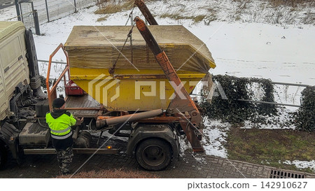 A worker operating a yellow skip loader truck to lift a covered waste container on a snowy winter day, highlighting industrial construction site logistics and professional waste removal A worker operating a yellow skip loader truck to lift a covered waste container on a snowy winter day, highlighting industrial construction site logistics and professional waste removal 142910627