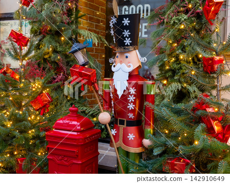 A festive nutcracker statue stands near a red post box and pine trees decorated with bright lights and presents, capturing the joyful atmosphere of a traditional outdoor holiday display A festive nutcracker statue stands near a red post box and pine trees decorated with bright lights and presents, capturing the joyful atmosphere of a traditional outdoor holiday display 142910649