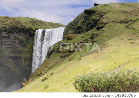 Skogafoss waterfall and sunlit grassy slopes with hillside steps in Iceland Skogafoss waterfall and sunlit grassy slopes with hillside steps in Iceland 142910769