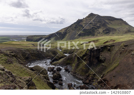 Skoga River Rapids and Grassy Gorge Beneath a Steep Mountain in Iceland Skoga River Rapids and Grassy Gorge Beneath a Steep Mountain in Iceland 142910807