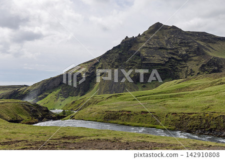 Skoga River and grassy banks beneath a rugged layered mountainside in Iceland Skoga River and grassy banks beneath a rugged layered mountainside in Iceland 142910808