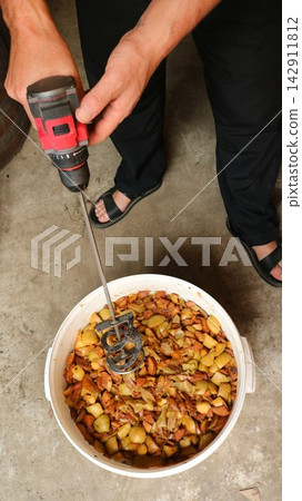 A person is holding a drill fitted with a special mixing attachment, using it to grind the apple pulp. A person is holding a drill fitted with a special mixing attachment, using it to grind the apple pulp. 142911812