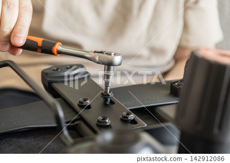 A hand uses a ratchet tool to tighten a bolt on a black metal office chair frame, demonstrating assembly work and furniture maintenance tasks within a home or professional setting A hand uses a ratchet tool to tighten a bolt on a black metal office chair frame, demonstrating assembly work and furniture maintenance tasks within a home or professional setting 142912086