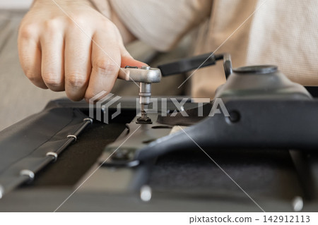 A hand uses a metal socket wrench to tighten a bolt on a black office chair seat base, demonstrating DIY furniture assembly and home maintenance tasks in a bright indoor setting A hand uses a metal socket wrench to tighten a bolt on a black office chair seat base, demonstrating DIY furniture assembly and home maintenance tasks in a bright indoor setting 142912113
