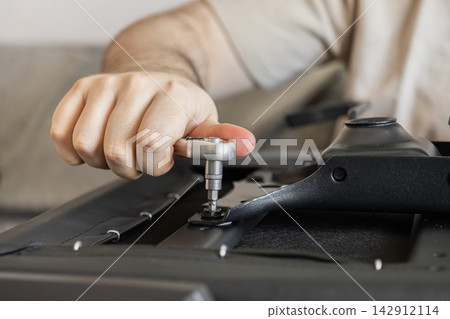 A man uses a metal ratchet tool to tighten a screw on the frame of an office chair, emphasizing DIY assembly and home improvement tasks within a residential interior living space A man uses a metal ratchet tool to tighten a screw on the frame of an office chair, emphasizing DIY assembly and home improvement tasks within a residential interior living space 142912114