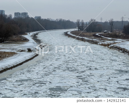 A frozen river flows through a snow-covered urban landscape on a chilly winter day, capturing the serene stillness and cold climate of the outdoors during the middle of the season A frozen river flows through a snow-covered urban landscape on a chilly winter day, capturing the serene stillness and cold climate of the outdoors during the middle of the season 142912141