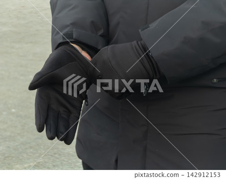 A person pulls on black winter gloves while wearing a matching warm jacket, conveying preparation for cold weather conditions and outdoor comfort during the chilly winter season A person pulls on black winter gloves while wearing a matching warm jacket, conveying preparation for cold weather conditions and outdoor comfort during the chilly winter season 142912153