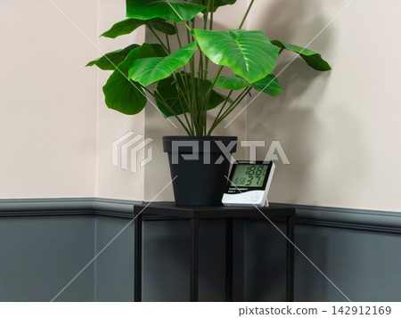 An indoor climate monitor rests on a side table with a large green plant, highlighting modern technology for checking ambient temperature and humidity levels within a cozy interior An indoor climate monitor rests on a side table with a large green plant, highlighting modern technology for checking ambient temperature and humidity levels within a cozy interior 142912169