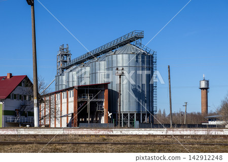 Industrial grain storage silos made of corrugated metal sit near a railway track and a brick tower under a clear blue sky, representing modern agricultural infrastructure and trade Industrial grain storage silos made of corrugated metal sit near a railway track and a brick tower under a clear blue sky, representing modern agricultural infrastructure and trade 142912248