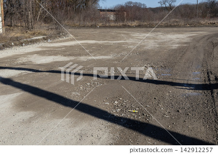 A desolate rural dirt road with scattered gravel and puddles stretches towards a distant thicket, emphasizing a sense of neglect and the harsh reality of unpaved infrastructure paths A desolate rural dirt road with scattered gravel and puddles stretches towards a distant thicket, emphasizing a sense of neglect and the harsh reality of unpaved infrastructure paths 142912257