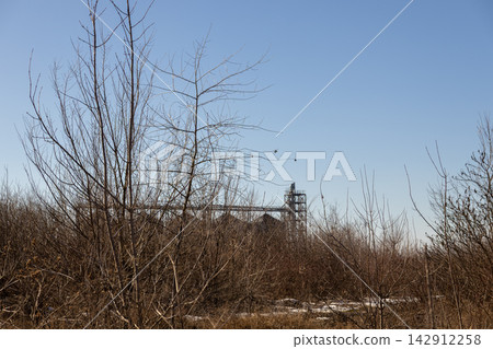 A tall industrial grain silo facility rises above barren winter brush and leafless trees against a clear blue sky, capturing the stark contrast of agriculture and nature in cold 142912258