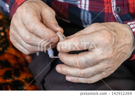 An elderly person holds a small hearing aid device in wrinkled hands, representing accessibility and improved quality of life for seniors managing natural age-related sensory changes An elderly person holds a small hearing aid device in wrinkled hands, representing accessibility and improved quality of life for seniors managing natural age-related sensory changes 142912269