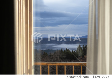 A scenic mountain landscape viewed from a window with sheer curtains, showing misty evergreen forests and rolling hills during a tranquil dawn in a peaceful rural travel destination 142912273