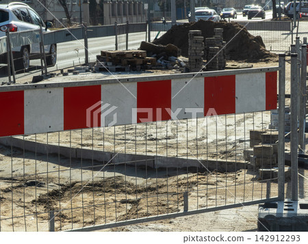 A red and white striped metal fence surrounds a dusty urban construction site where road paving materials are staged beside a busy city street, indicating zone safety and maintenance A red and white striped metal fence surrounds a dusty urban construction site where road paving materials are staged beside a busy city street, indicating zone safety and maintenance 142912293