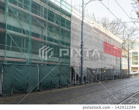 A building facade covered in scaffolding and safety mesh, standing along a paved street with tram rails, representing an urban renovation project in a city center architectural setting A building facade covered in scaffolding and safety mesh, standing along a paved street with tram rails, representing an urban renovation project in a city center architectural setting 142912294