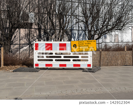 A red and white traffic safety barrier with a detour sign stands on a city sidewalk during the day, indicating construction work, urban maintenance, or restricted pedestrian paths A red and white traffic safety barrier with a detour sign stands on a city sidewalk during the day, indicating construction work, urban maintenance, or restricted pedestrian paths 142912304