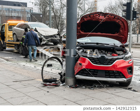 A damaged red car sits crashed against a metal pole at a city intersection, as a worker prepares to load a broken silver vehicle onto a yellow tow truck after a roadside accident event A damaged red car sits crashed against a metal pole at a city intersection, as a worker prepares to load a broken silver vehicle onto a yellow tow truck after a roadside accident event 142912315