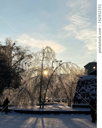 Sunlight filters through ice-covered trees in a frosty urban park at sunset, evoking a serene winter landscape with a glowing horizon and dramatic cold weather seasonal atmosphere Sunlight filters through ice-covered trees in a frosty urban park at sunset, evoking a serene winter landscape with a glowing horizon and dramatic cold weather seasonal atmosphere 142912331