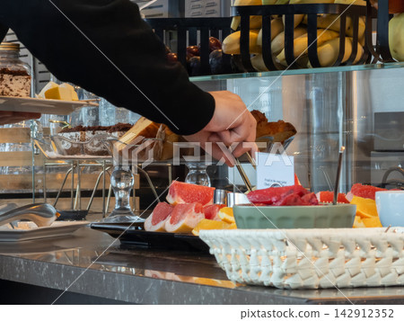 A guest hand uses tongs for fresh grapefruit slices at a hotel breakfast buffet with assorted fruits and baked goods, capturing a morning hospitality dining experience in a restaurant A guest hand uses tongs for fresh grapefruit slices at a hotel breakfast buffet with assorted fruits and baked goods, capturing a morning hospitality dining experience in a restaurant 142912352